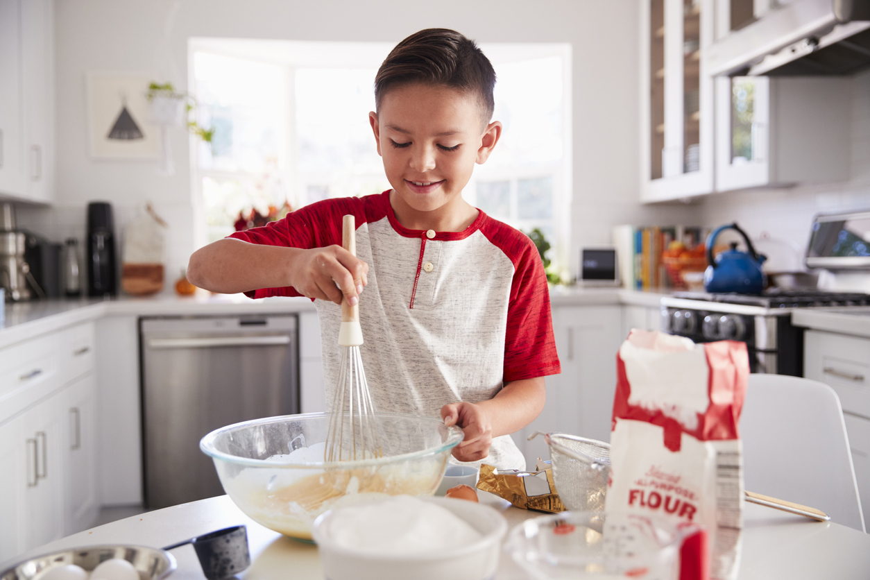 A young child in a kitchen mixing dough in a bowl with various cooking elements on the table
around him such as flour, eggs, and sugar